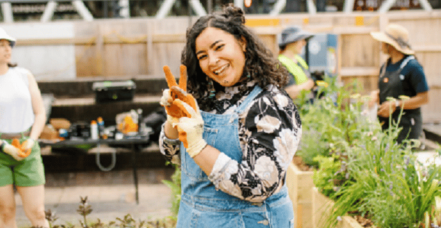 A women with gardening gloves on making the peace sign in a community garden area showcasing social value.