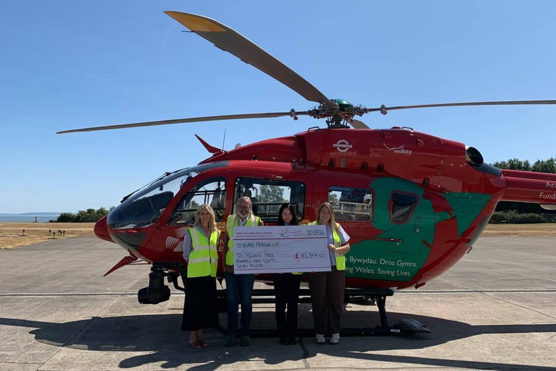 Abi Pearce, Lee Fisher, Menglin Li and Isabelle Mann holding a cheque totally the amount raised for the Wales Air Ambulance, in front of a helicopter.